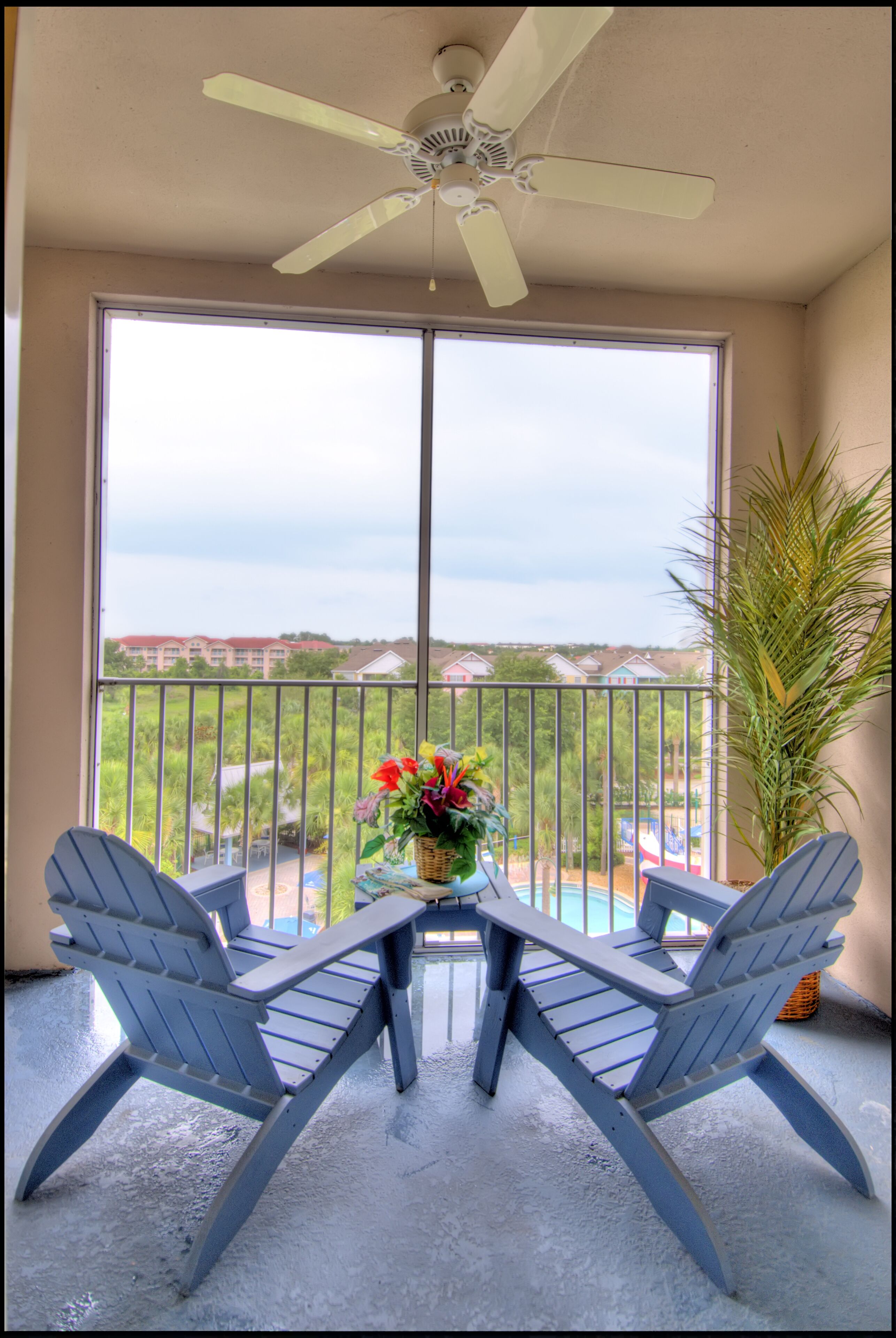 A balcony with two Adirondack chairs, a ceiling fan, and a view of a swimming pool and distant buildings.