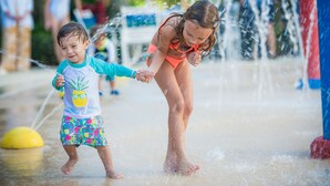 Un niño y un adulto jugando en una zona de juegos acuáticos, con una pelota amarilla y chorros de agua.