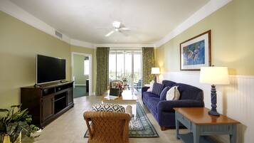 A living room with a flat-screen TV, a sofa, a coffee table, a wicker chair, a lamp, and a painting on the wall.