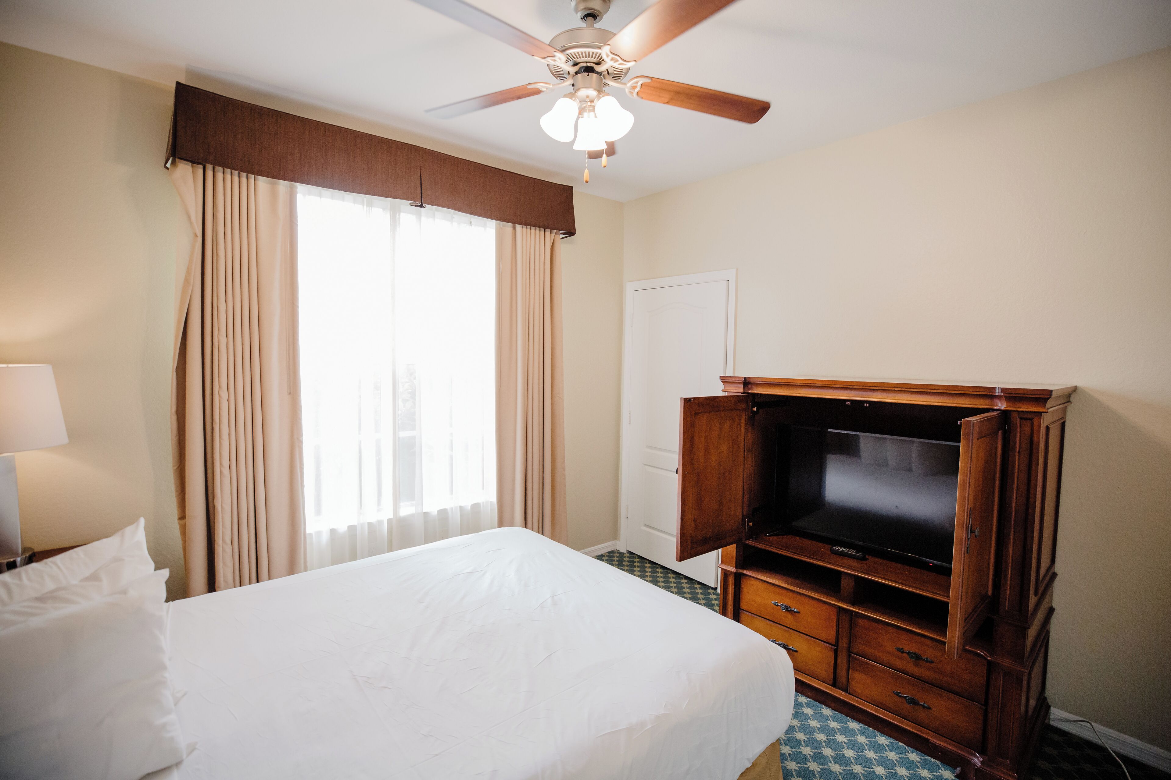 Una habitación de hotel con una cama, televisión, ventilador de techo y ventana con cortinas.