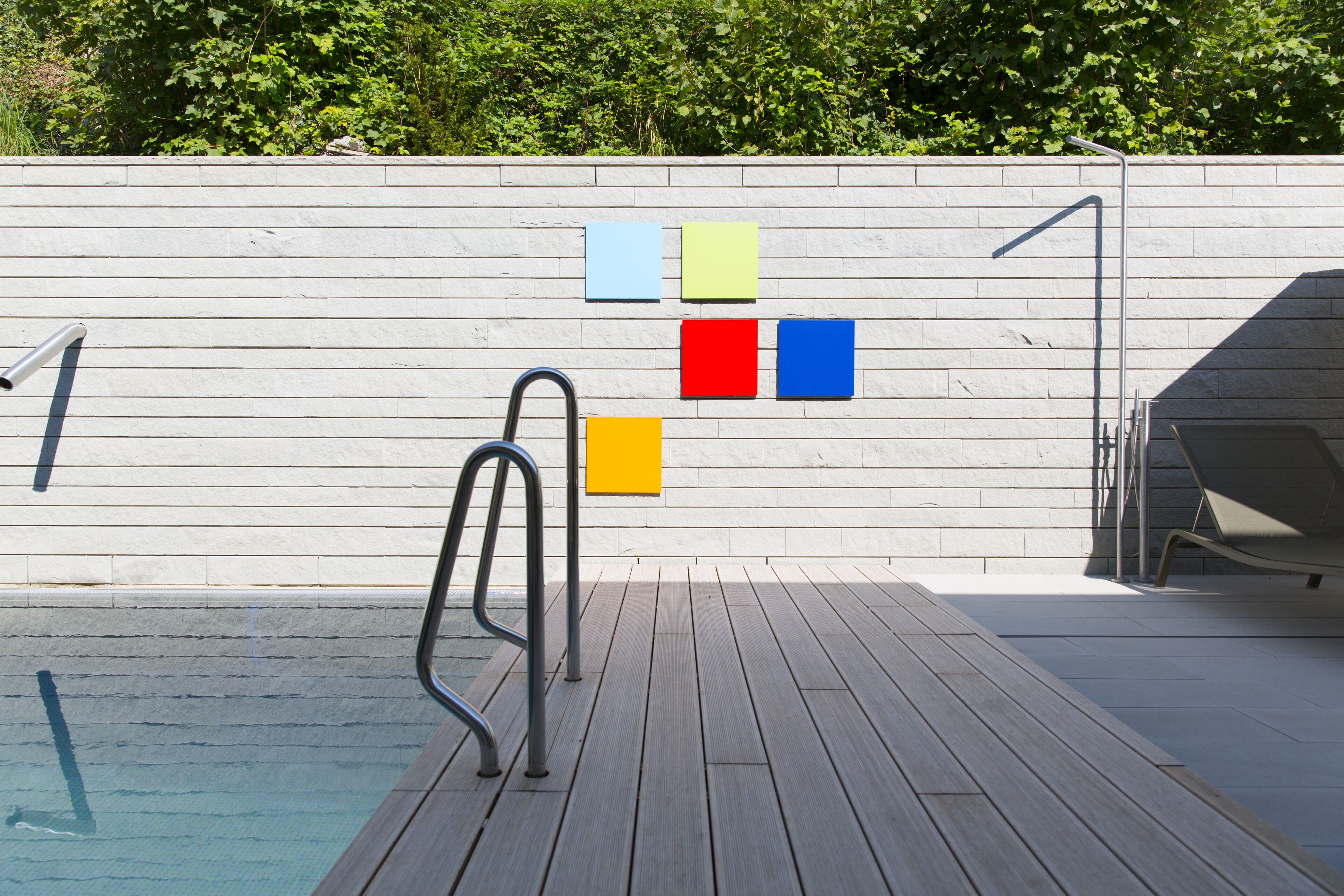 Un espace piscine doté d’une terrasse en bois, d’une échelle en métal et d’un mur orné de motifs carrés aux couleurs vives.