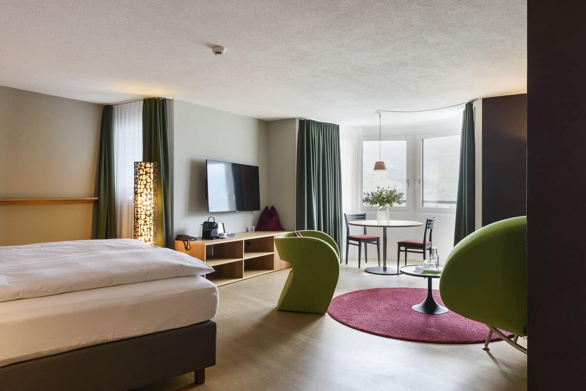 A hotel room with a bed, a TV, a desk with a chair, a dining table with chairs, and a window with curtains.