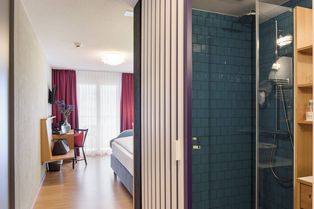 A hotel room with a bed, a desk with a chair, a desk lamp, a vase with flowers, and a shower area with blue tiles.