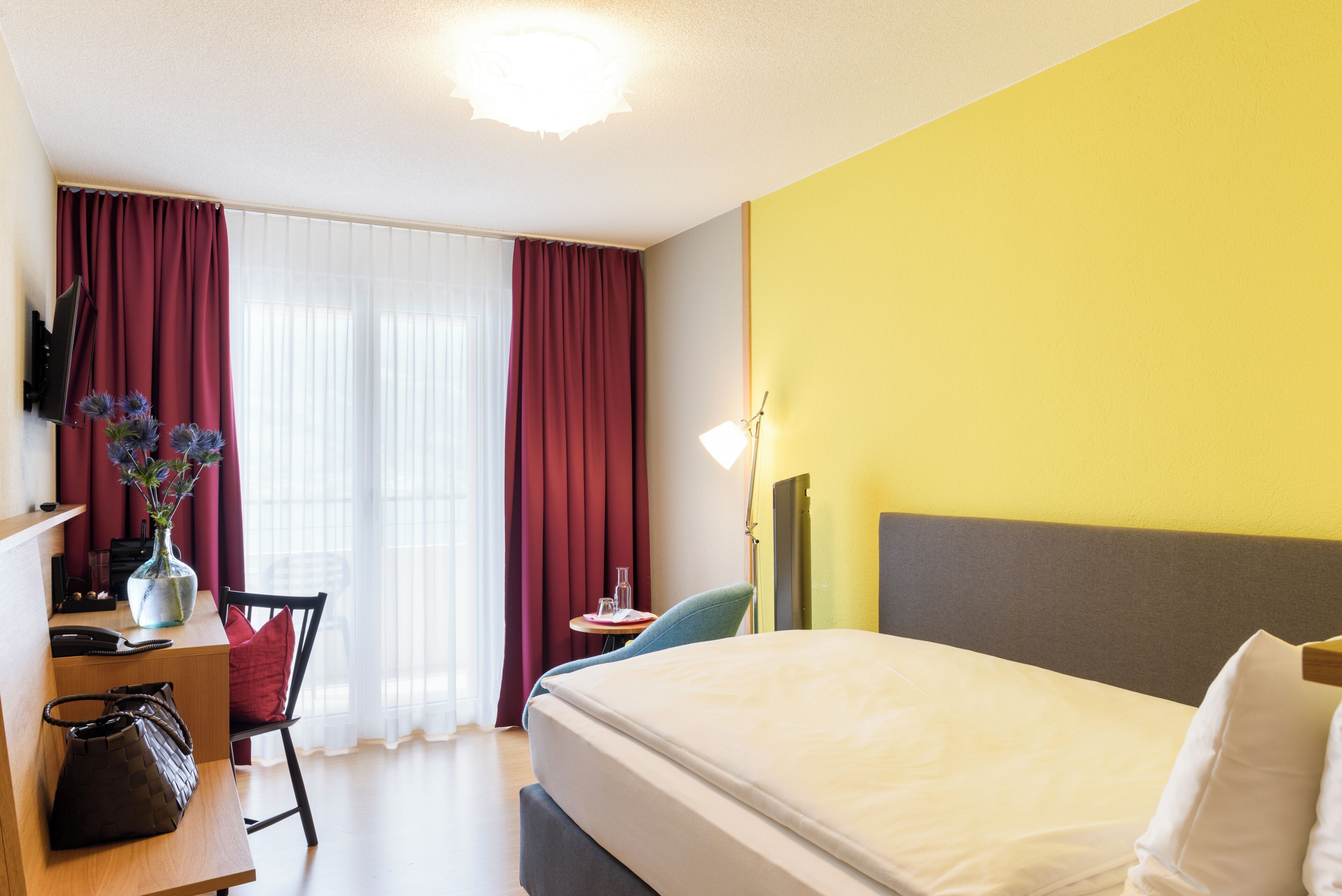 Una habitación de hotel con una cama, un escritorio con silla, una lámpara y un florero con flores.
