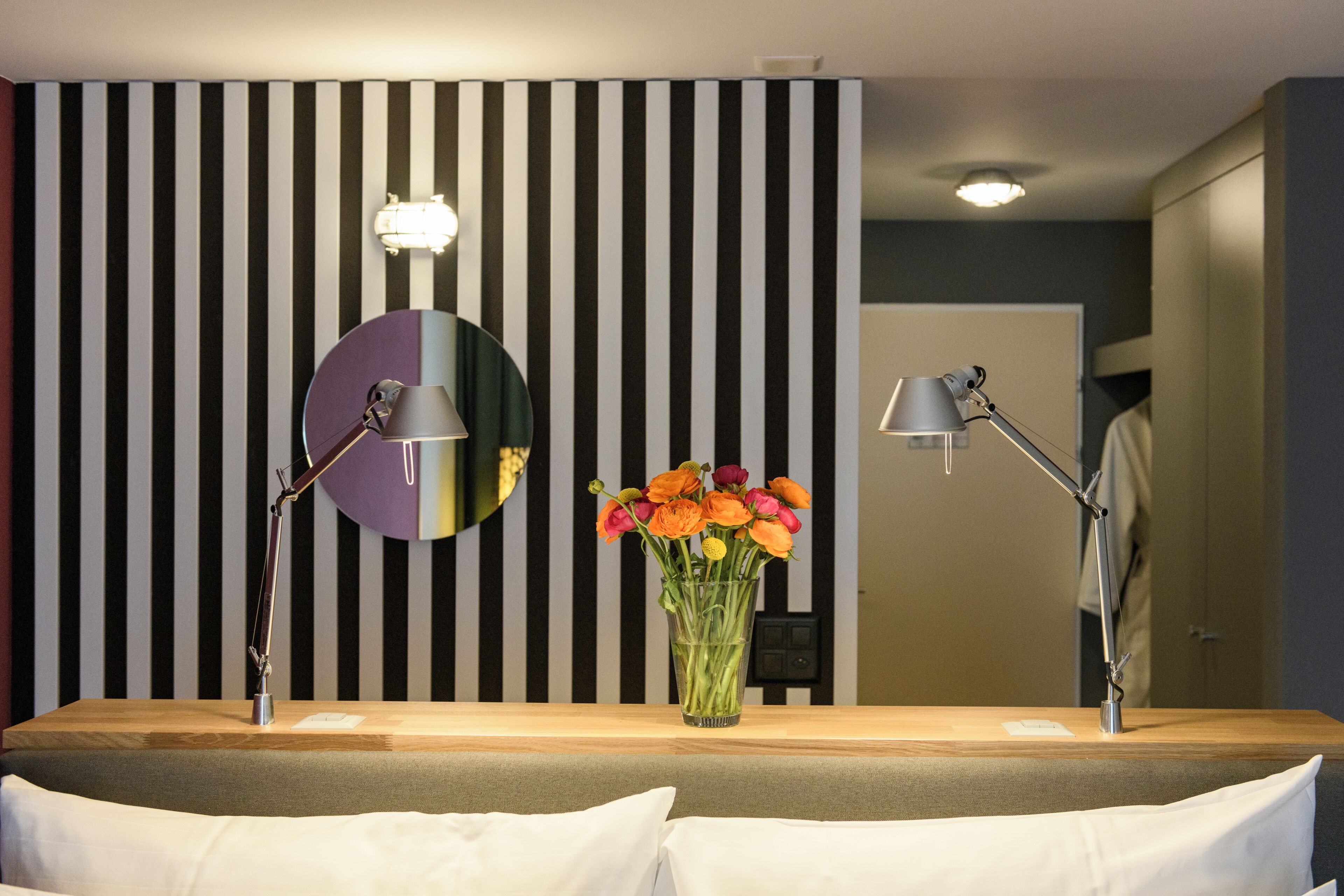 Een hotelkamer met een gestreepte wand, een ronde spiegel, twee bedlampen en een bloemenvaas.