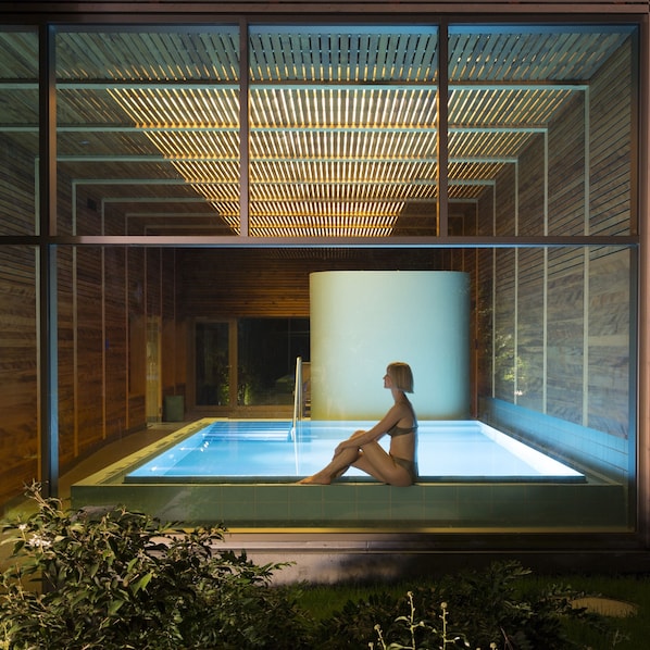 A modern indoor pool with a glass enclosure, a person sitting by the pool, and a wooden ceiling structure.