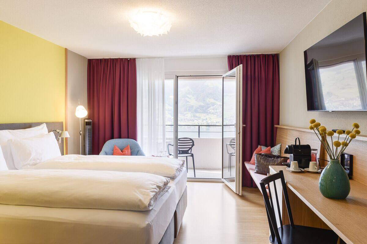 A hotel room with a bed, a desk with a chair, a TV, and a balcony with a view.