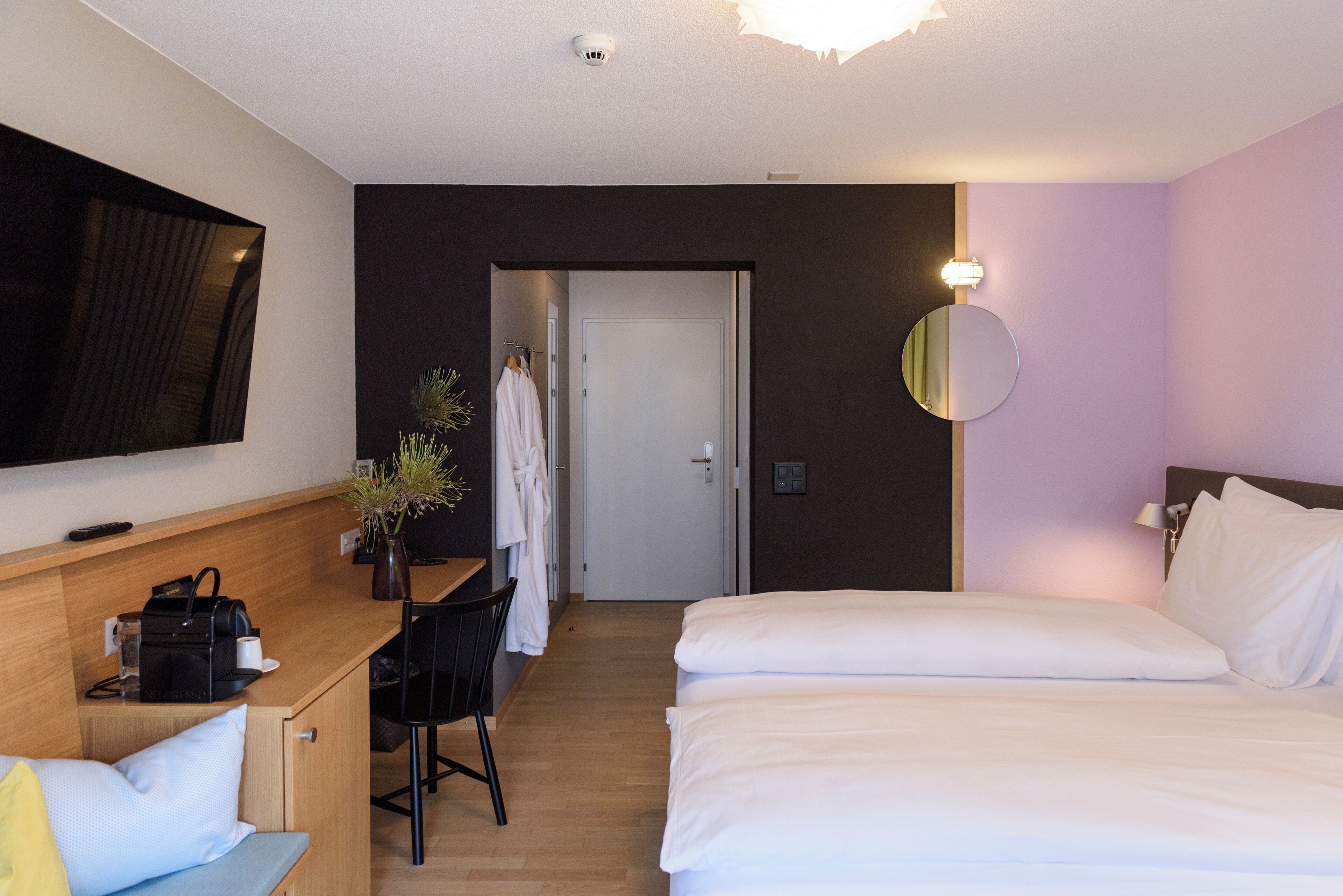 A hotel room with a bed, a desk with a chair, a TV mounted on the wall, a mirror, and a door leading to another room.