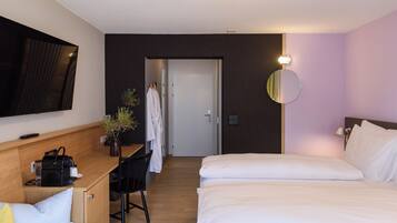 A hotel room with a bed, a desk with a chair, a TV mounted on the wall, a mirror, and a door leading to another room.