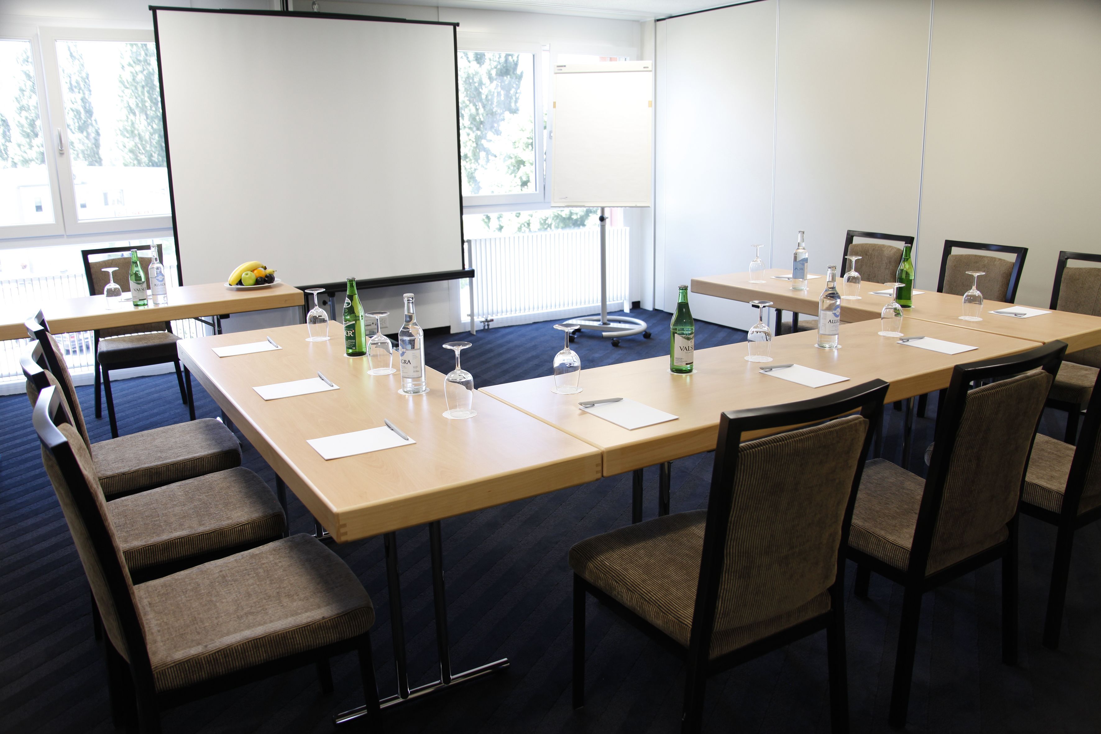 A conference room with a large table, chairs, a projector screen, and a window with a view of trees.