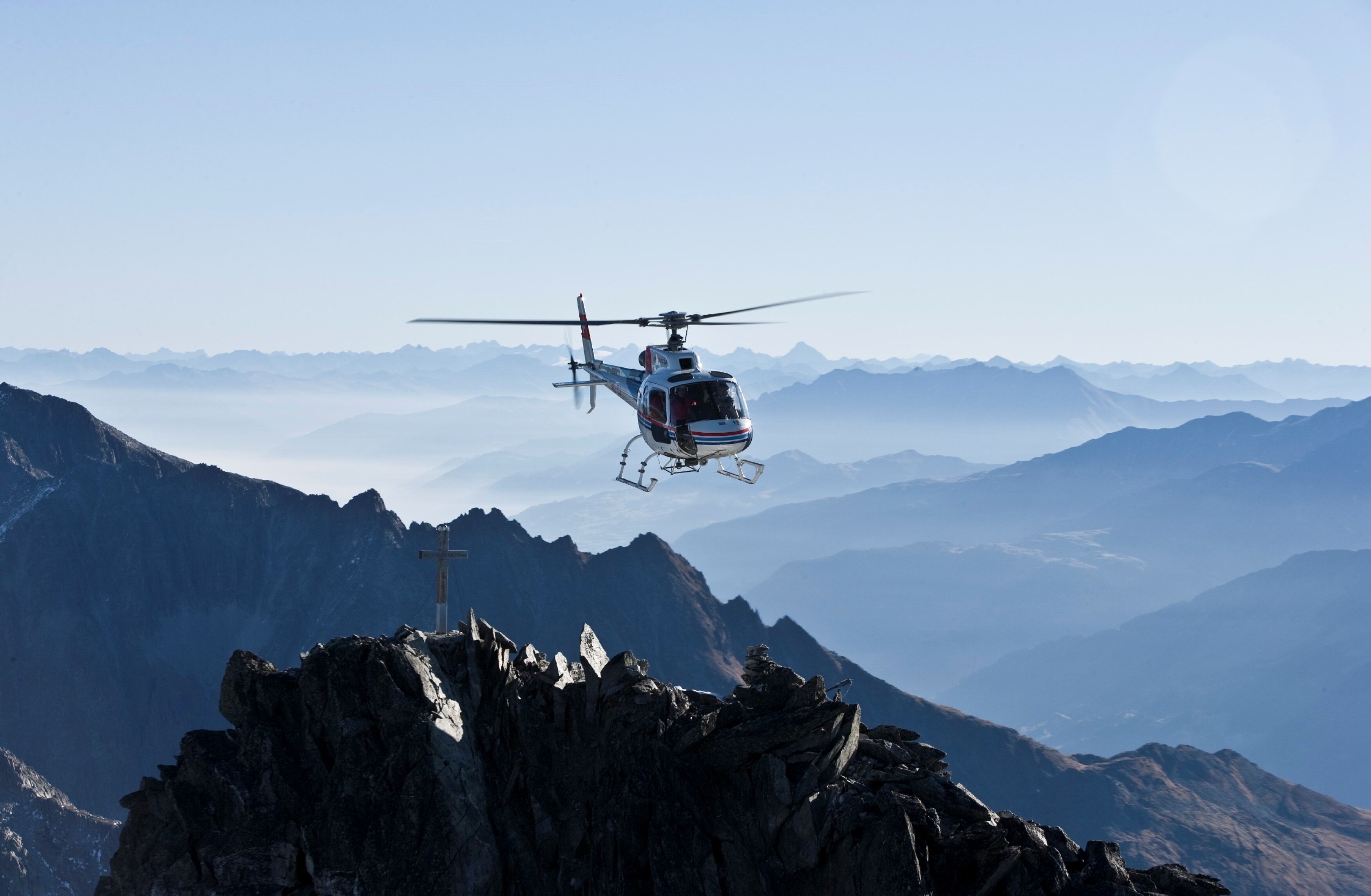 A helicopter flying over a mountainous landscape with rocky terrain and a cross on a peak.