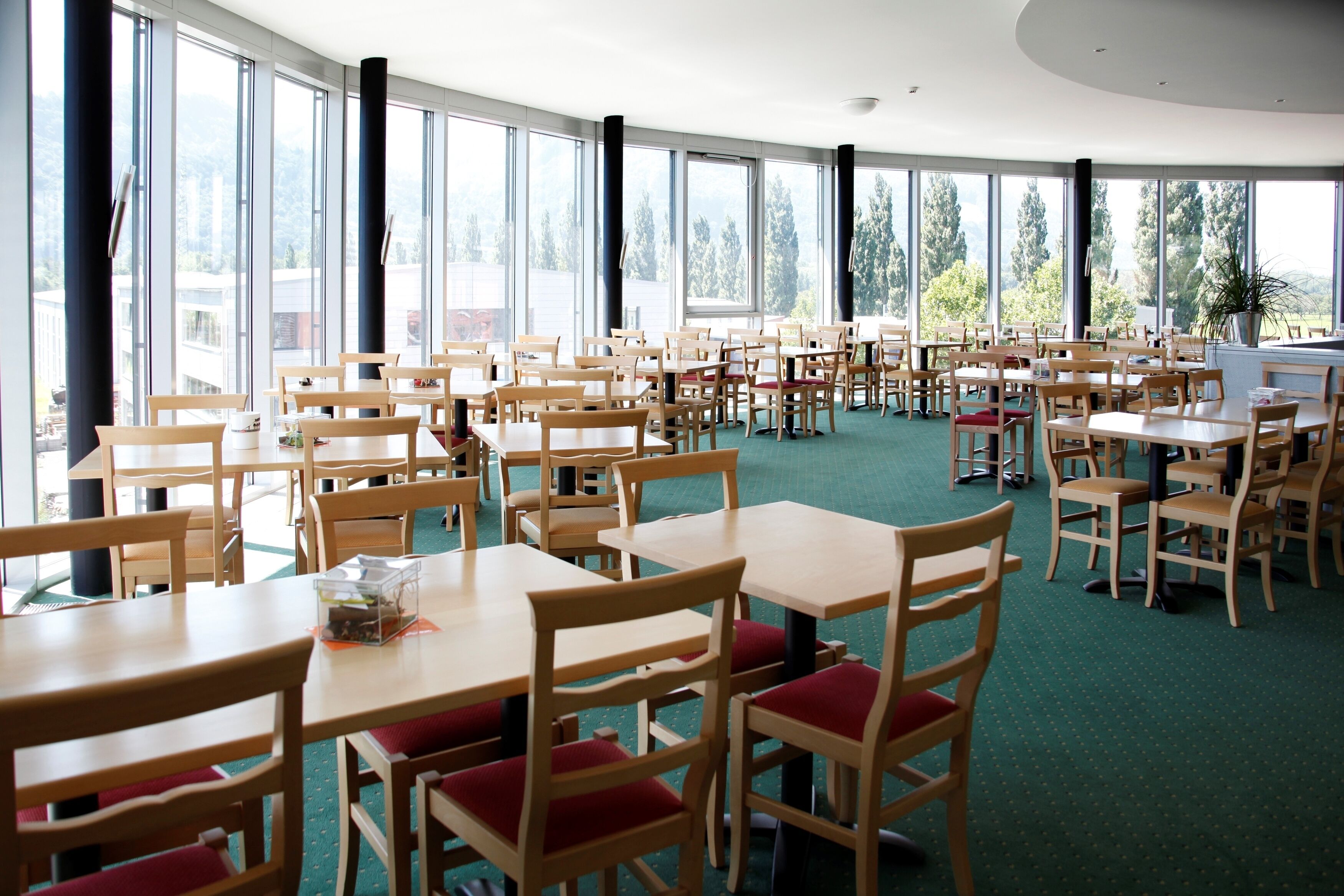 Un vaste espace dédié à la salle à manger, avec de nombreuses tables et chaises en bois, de grandes fenêtres offrant une vue sur l’extérieur et un sol recouvert d’une moquette verte.