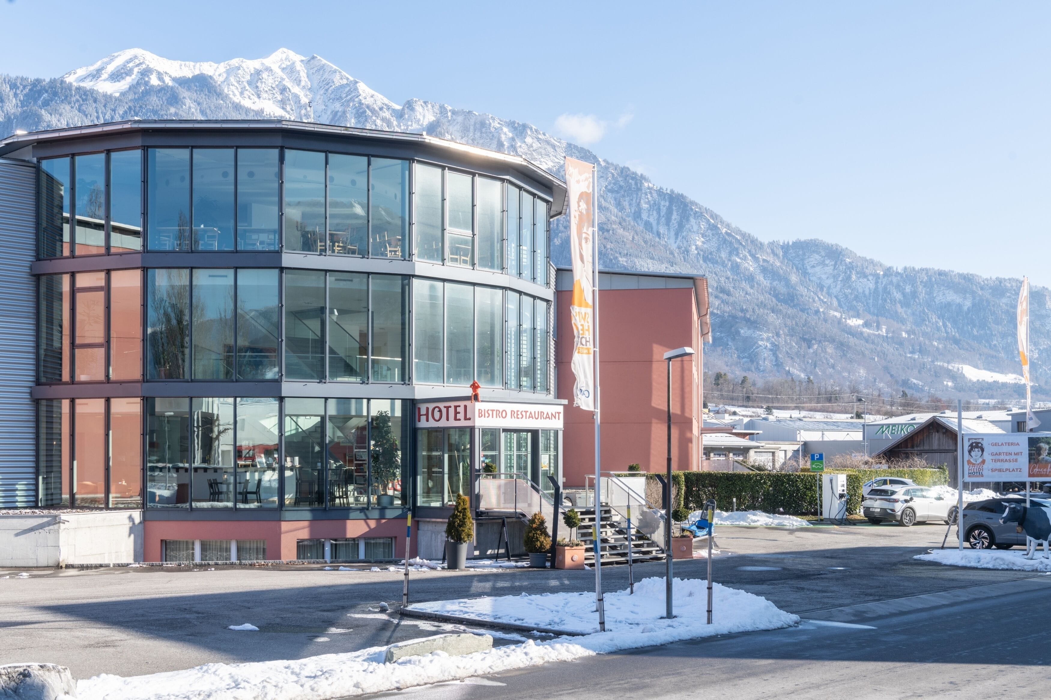 A modern hotel with a glass facade, a visible restaurant, and a parking area.