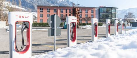 Una fila de estaciones de carga para vehículos eléctricos Tesla en un estacionamiento nevado, con un hotel y montañas al fondo.