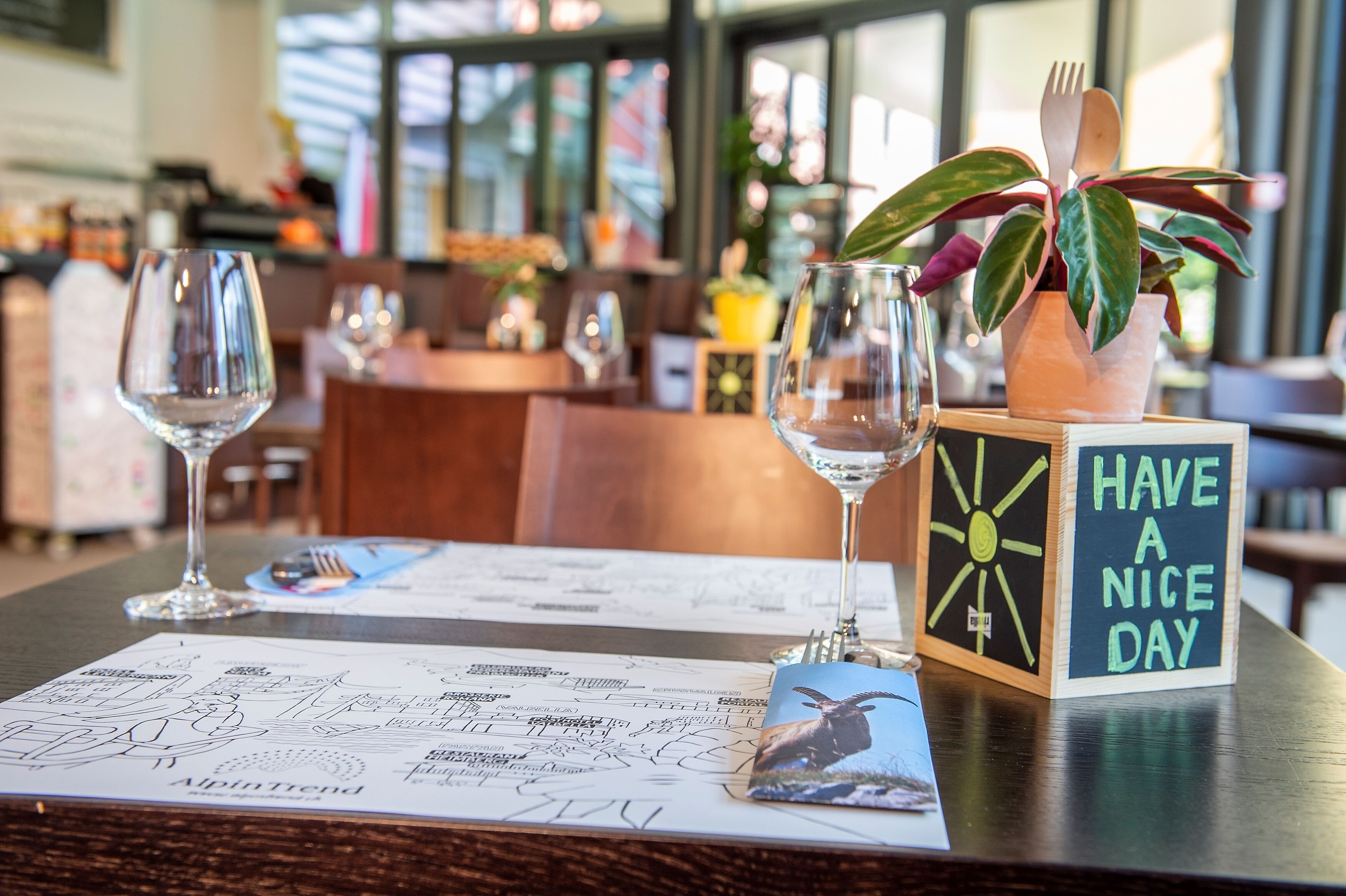 A restaurant table with a menu, a glass of water, a glass of wine, a wooden block with a message, and a potted plant with a fork and spoon.