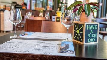 A restaurant table with a menu, a glass of water, a glass of wine, a wooden block with a message, and a potted plant with a fork and spoon.