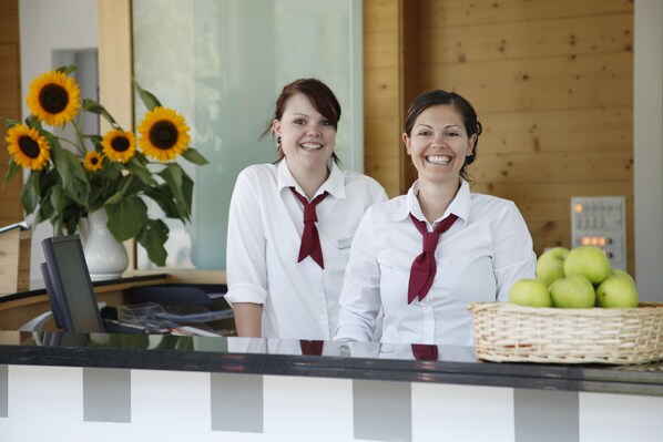 Two receptionists in white shirts with red ties standing behind a reception desk with a basket of green apples and a vase of sunflowers.