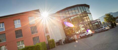 A hotel with a modern glass facade, a parking area with several cars, and a clear sky.