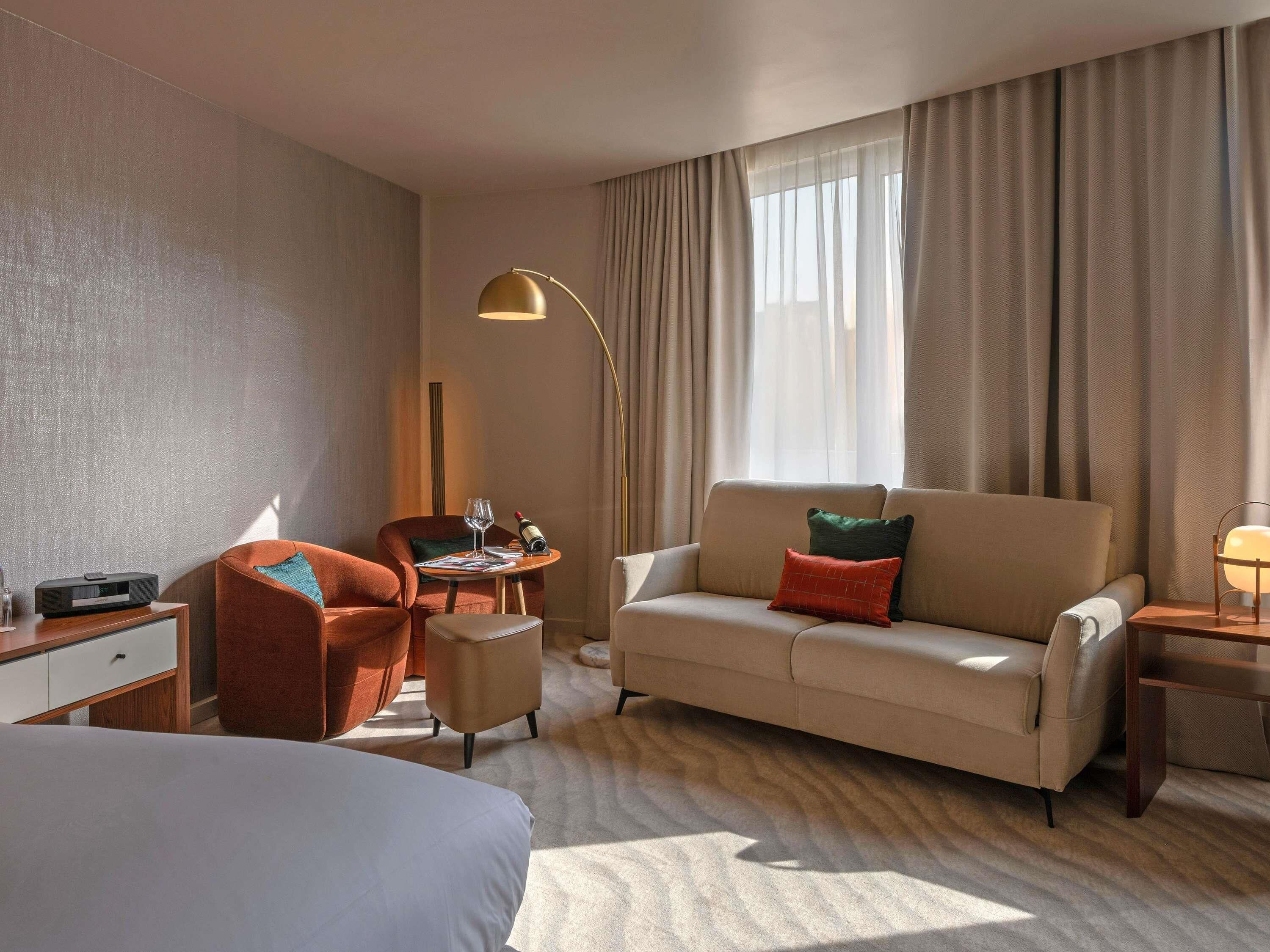 A hotel room with a bed, a sofa, a desk with a lamp, and a window with curtains.