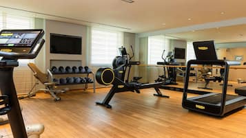 A gym with cardio machines, dumbbells, and a TV screen.