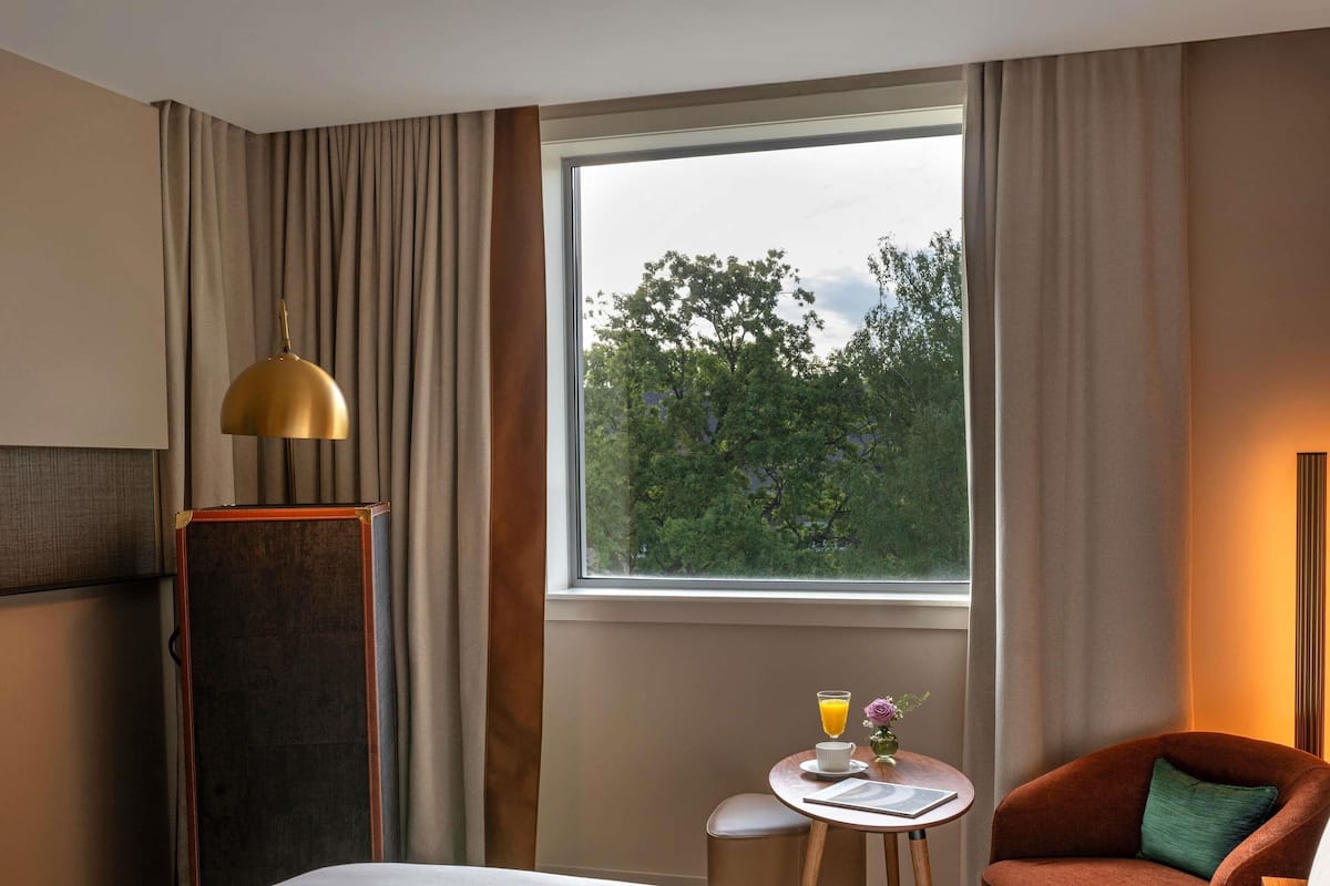 Ein Hotelzimmer mit einem Bett, einem kleinen runden Tisch mit Getränk und Blumenvase, einem Sessel und einem Fenster mit Blick auf Bäume.