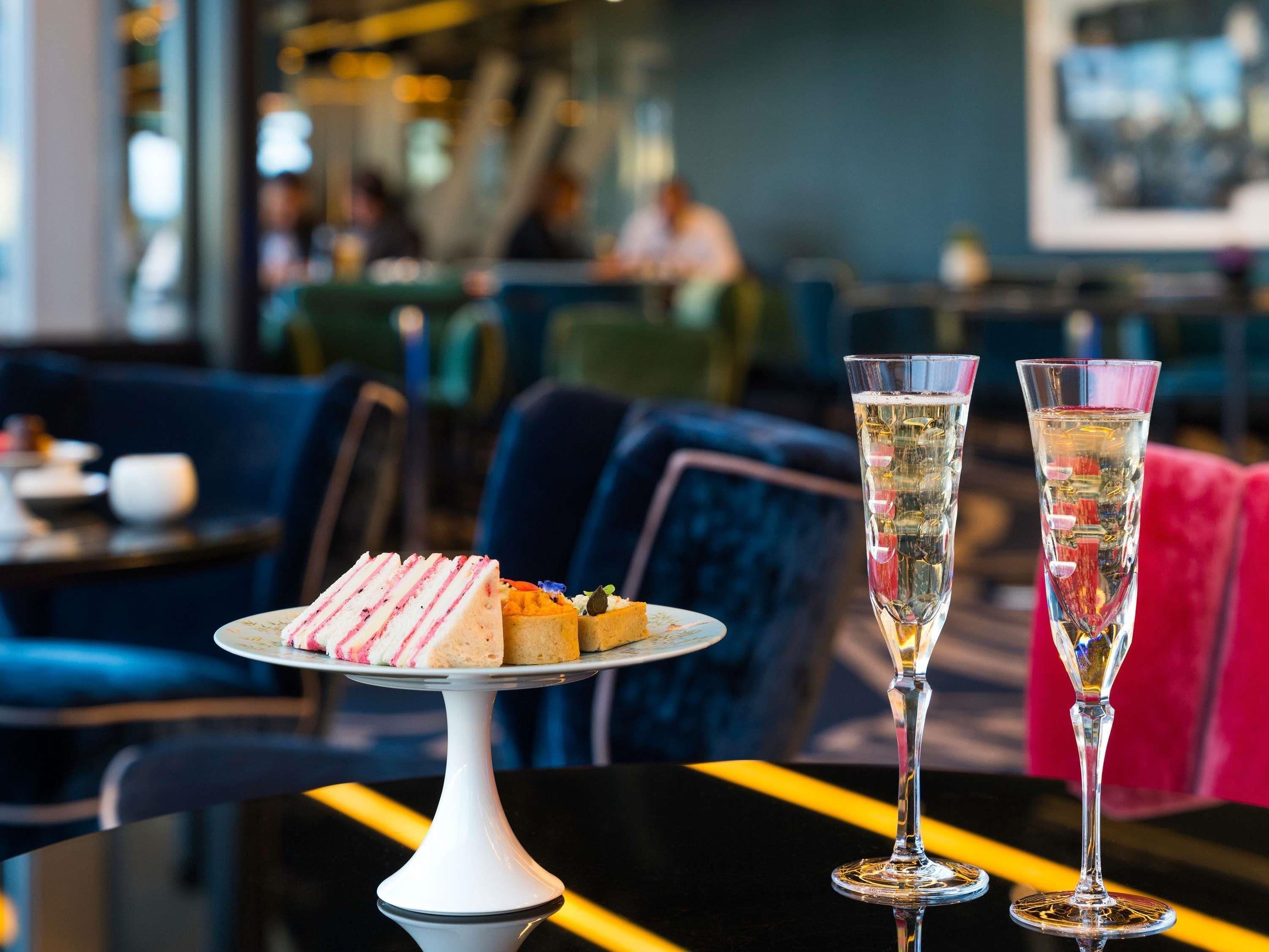 A dessert plate with various pastries on a white pedestal, two tall glasses of champagne, and a modern interior with seating.