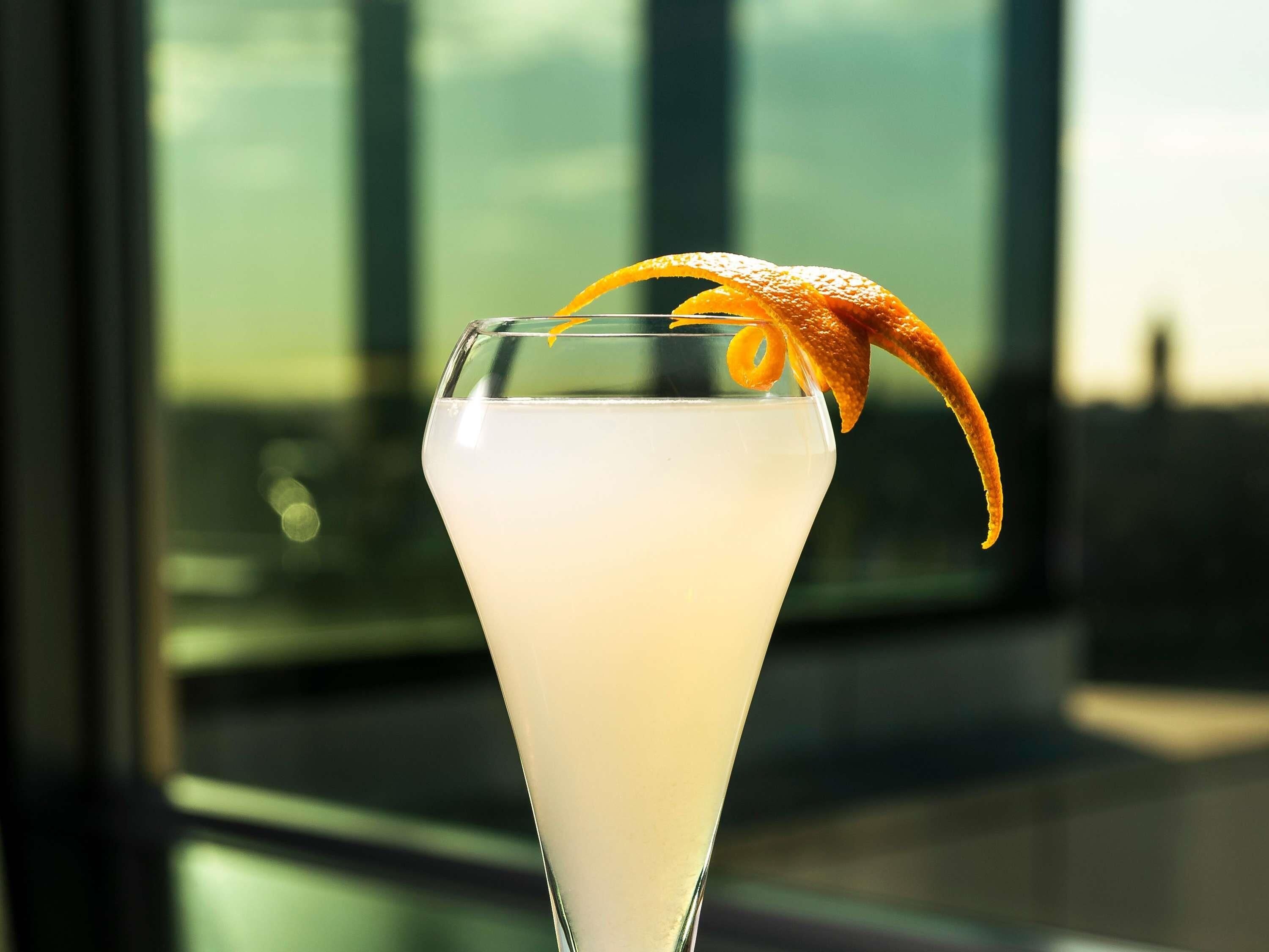 A cocktail in a tall glass with an orange peel garnish, set against a blurred indoor background with large windows.