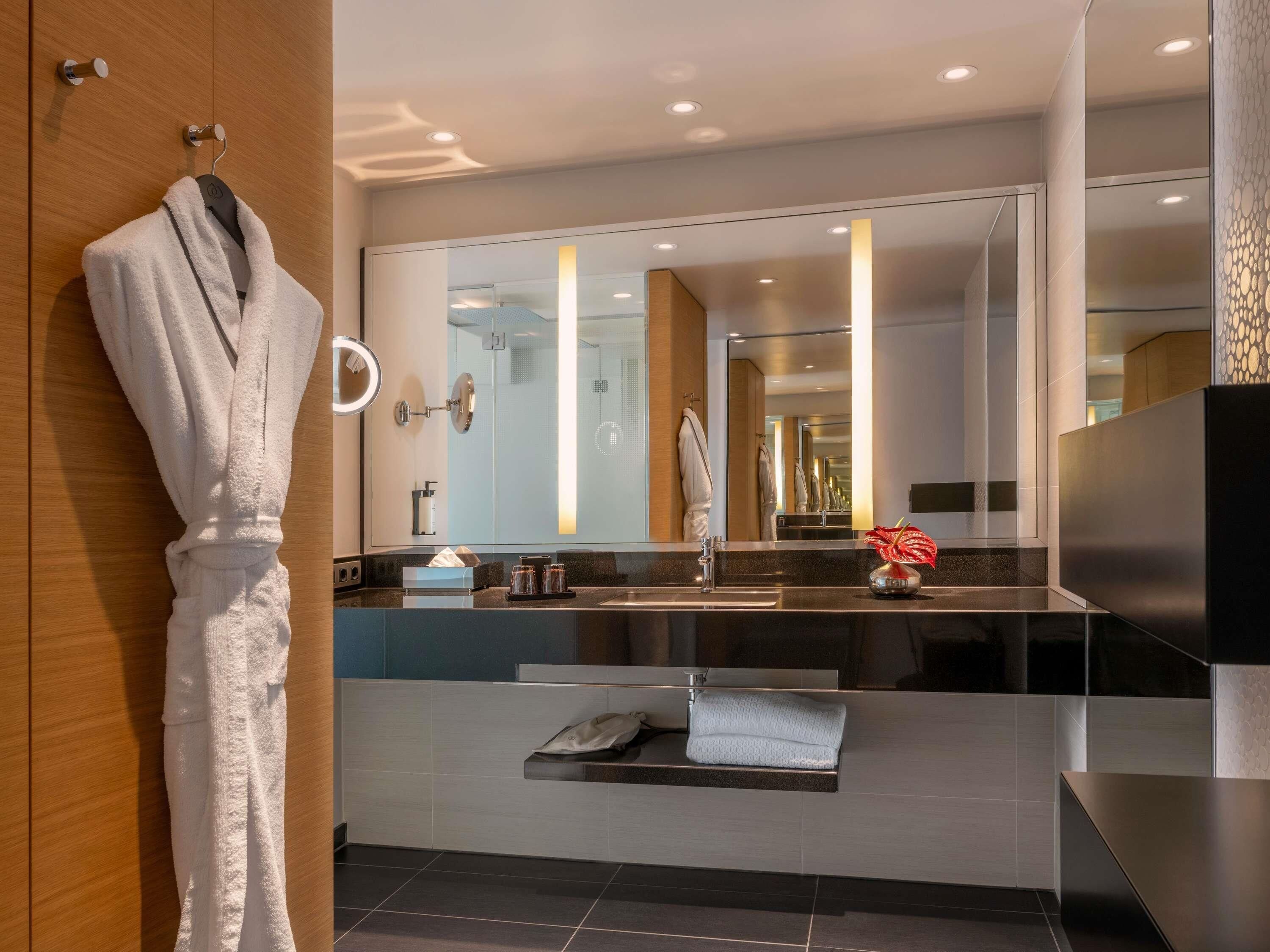 A modern bathroom with a large mirror, a robe hanging on a hook, a towel rack, a sink with a towel, and a small vase with a flower.