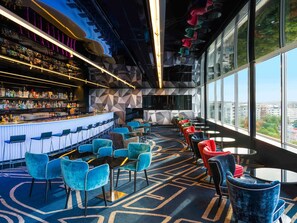 A modern bar area with a bar counter, seating, and large windows offering a city view.