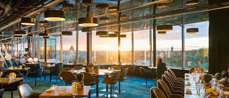 A modern restaurant with high ceilings, large windows, and a view of a cityscape at sunset.
