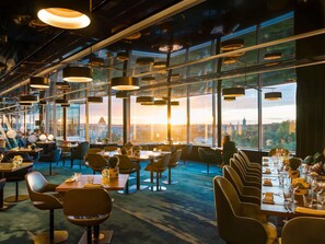 A modern restaurant with high ceilings, large windows, and a view of a cityscape at sunset. - Sofitel Luxembourg Le Grand Ducal (Luxembourg City)