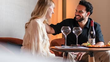 Un couple est assis à une table, devant deux verres à vin et une bouteille de vin, dans une pièce où se trouvent un canapé et une lampe.