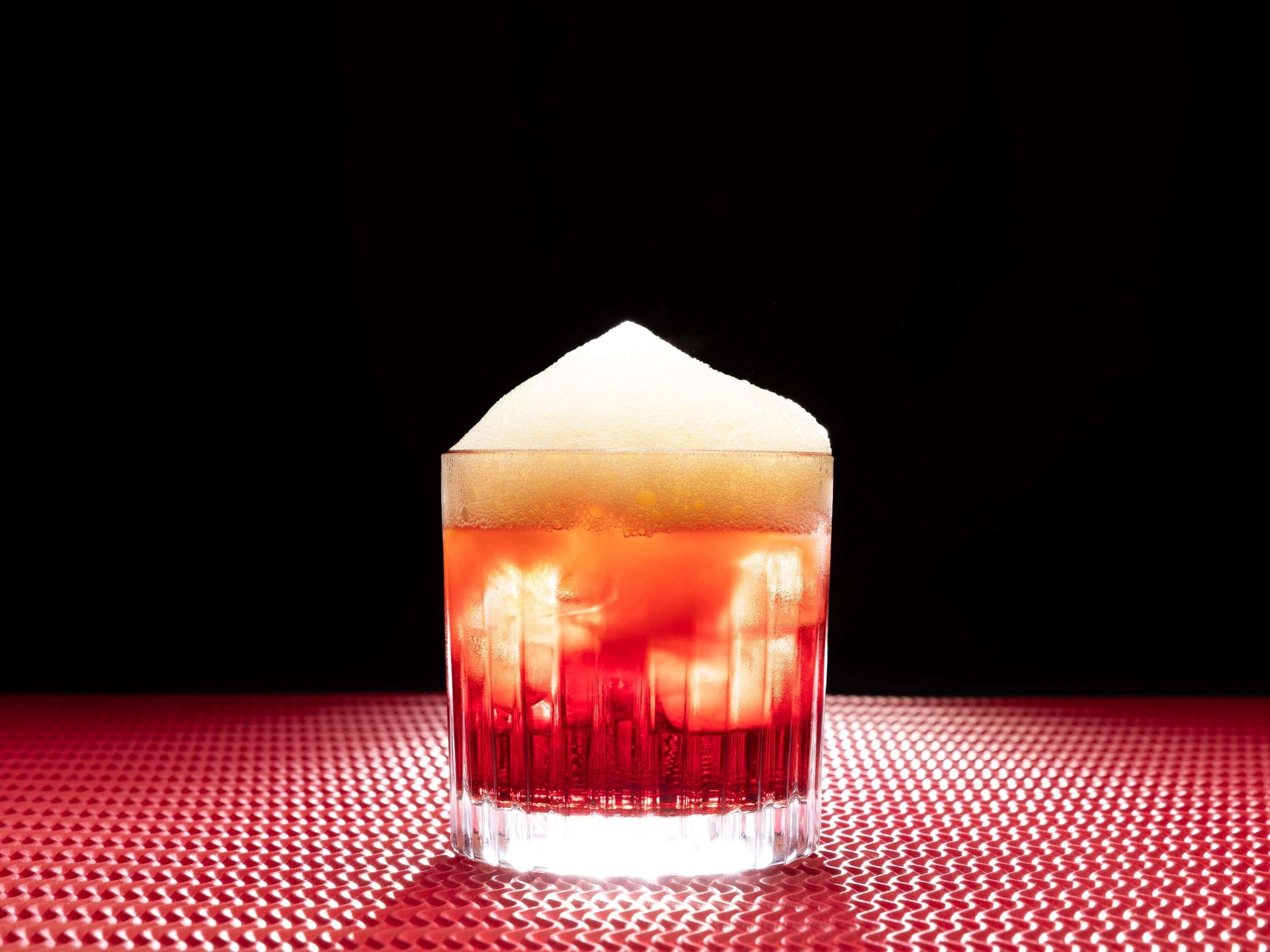 Un verre transparent contenant un liquide rouge et surmonté d’une mousse blanche, posé sur une surface rouge à texture marquée.
