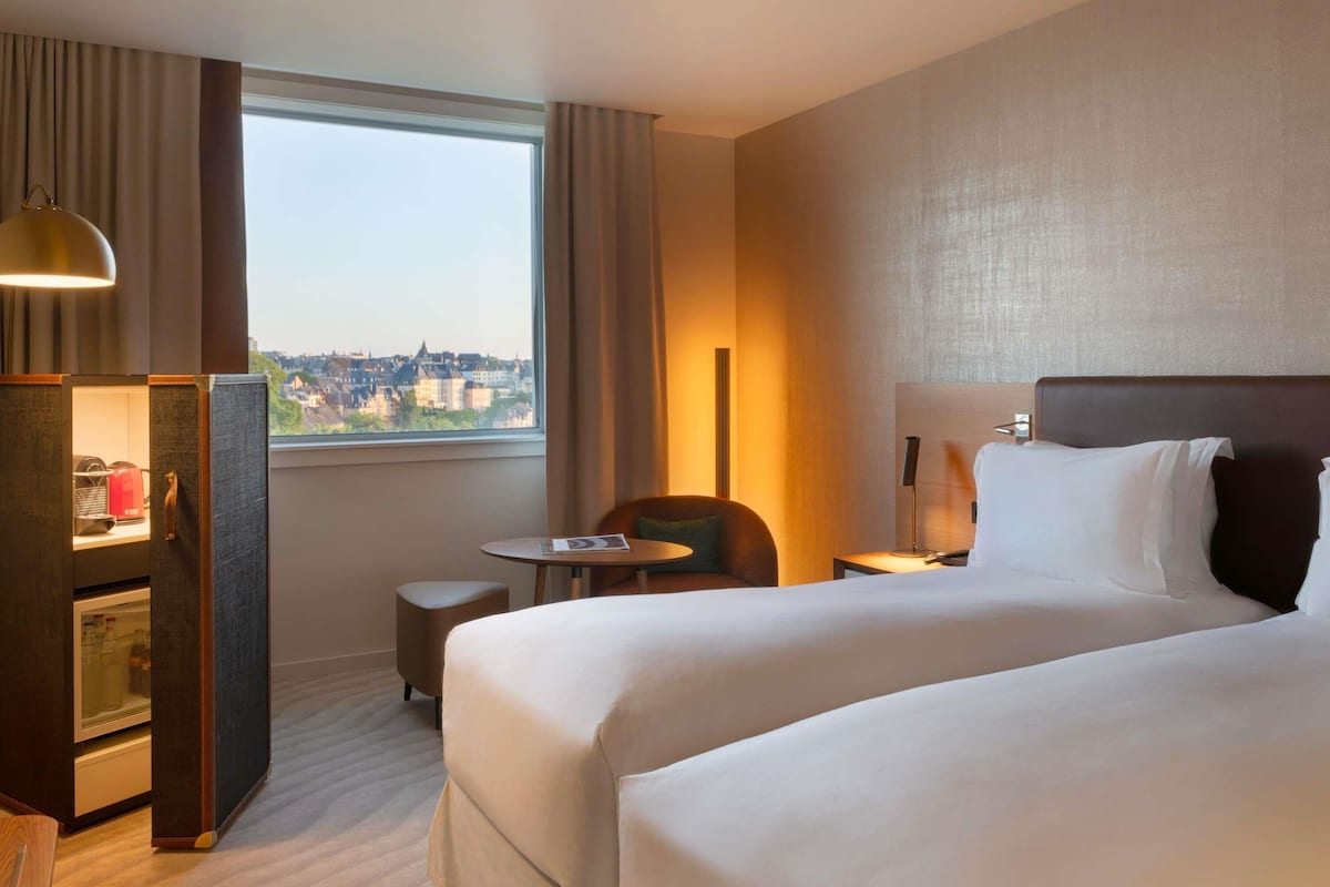 Ein Hotelzimmer mit Bett, Schreibtisch mit Lampe, Stuhl, kleinem Tisch und Blick auf eine Stadtlandschaft durch ein großes Fenster.