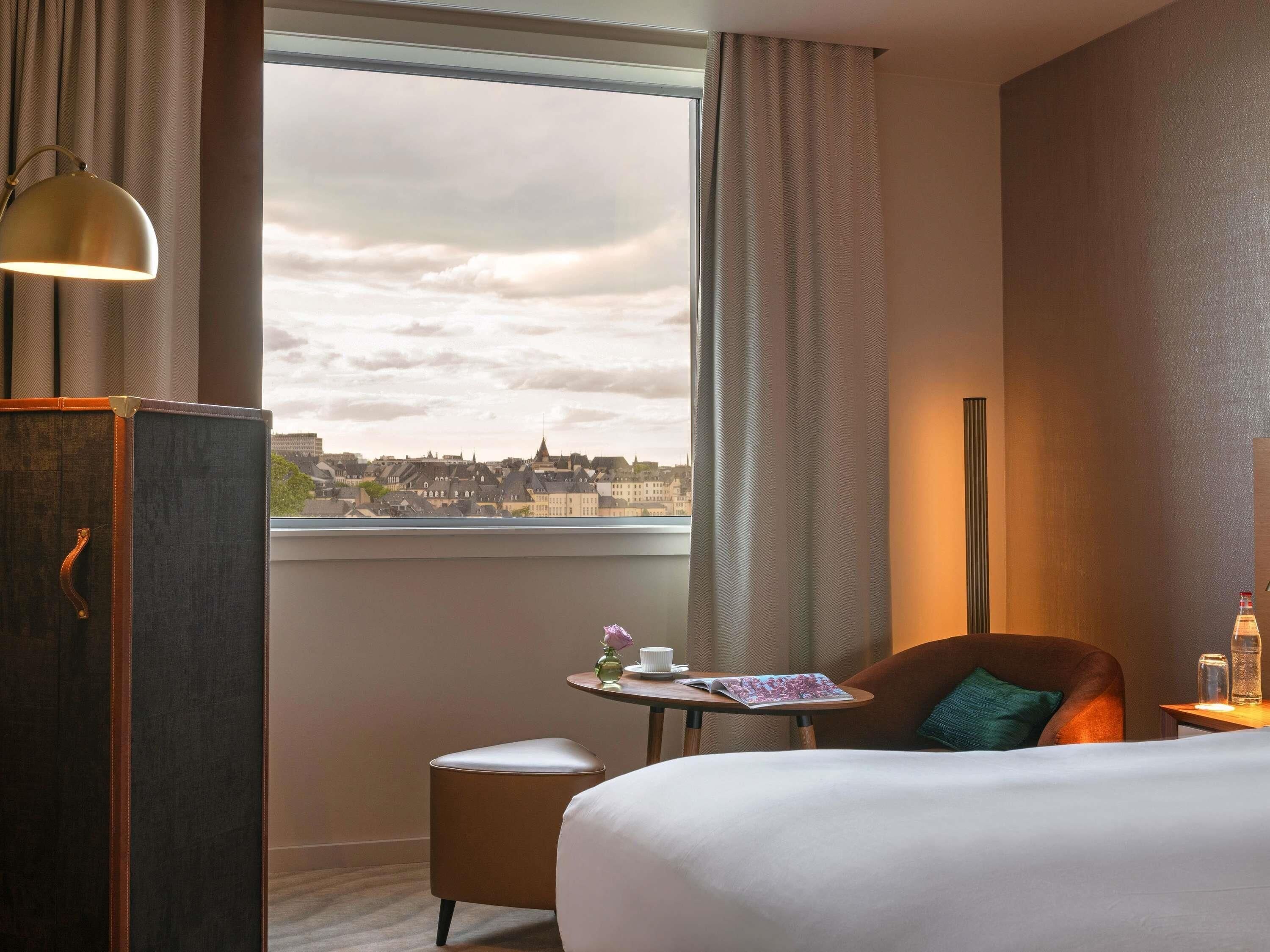 A hotel room with a bed, a small table with a cup and a book, a chair, and a view of a cityscape through a window.