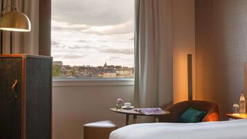 A hotel room with a bed, a small table with a cup and a book, a chair, and a view of a cityscape through a window.