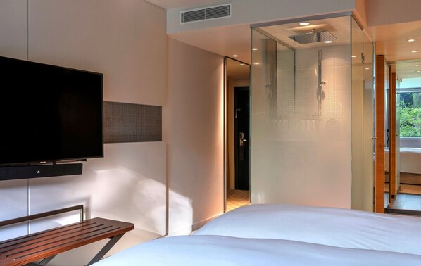 A modern hotel room with a large bed, a flat-screen TV mounted on the wall, and a glass shower enclosure.
