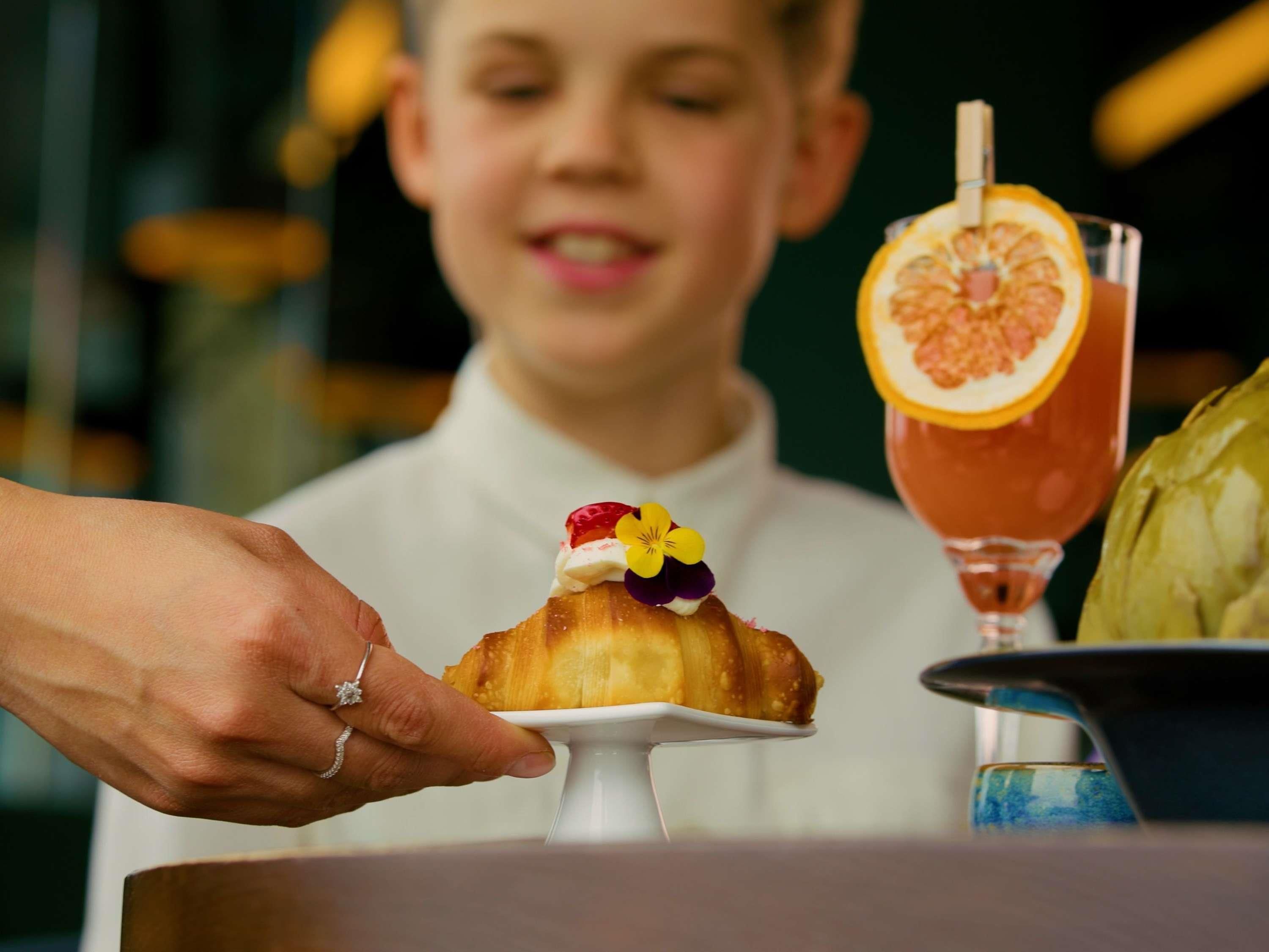 Una persona sostiene un pastel con cobertura decorativa, un vaso con guarnición de fruta y un alcachofa en un plato.