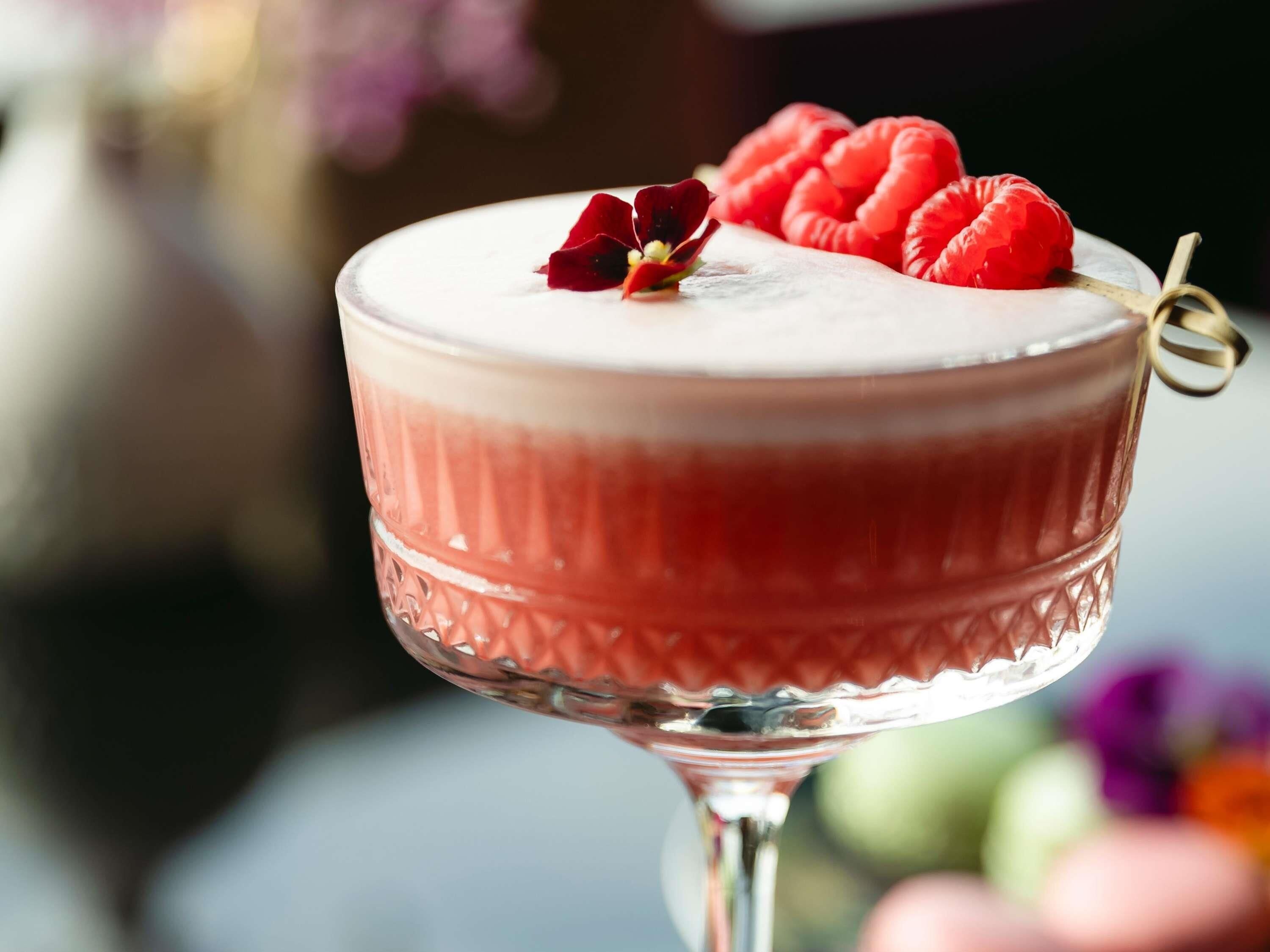A cocktail in a glass with a textured design, garnished with raspberries and a flower.