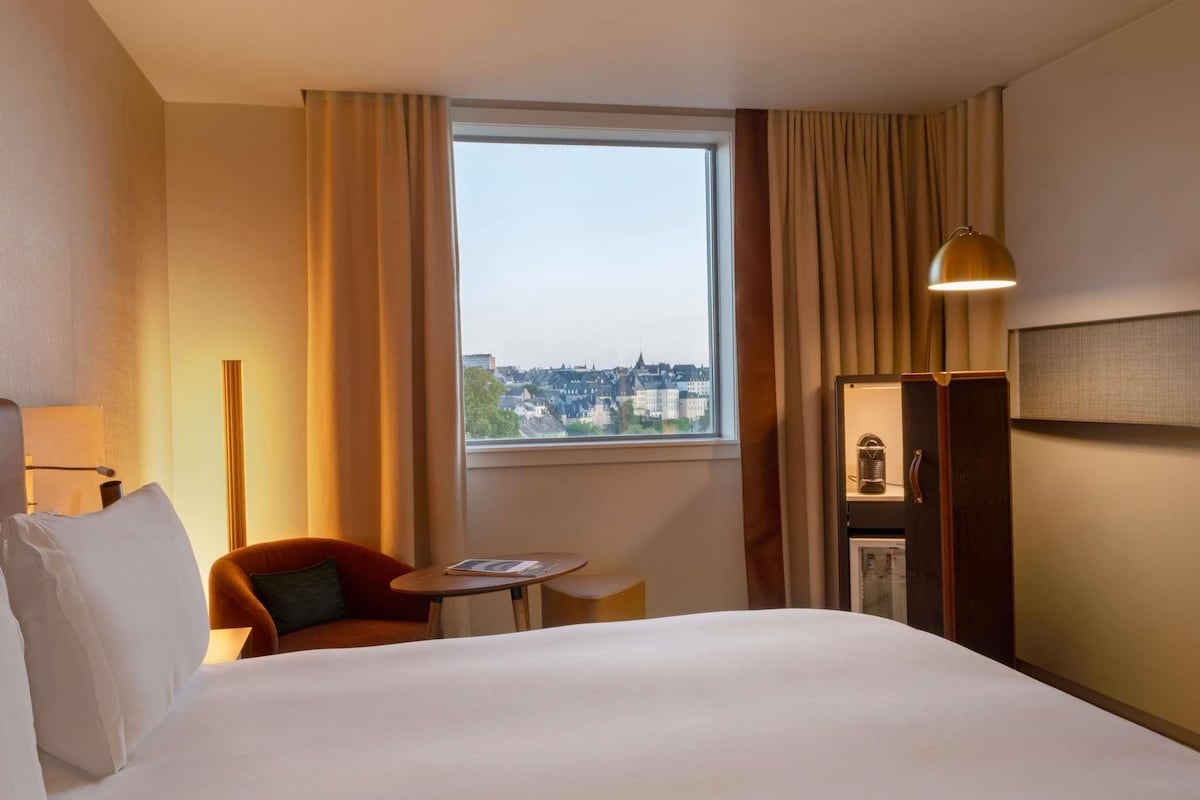 A hotel room with a bed, a chair, a small table, a lamp, and a view of a cityscape through a window.