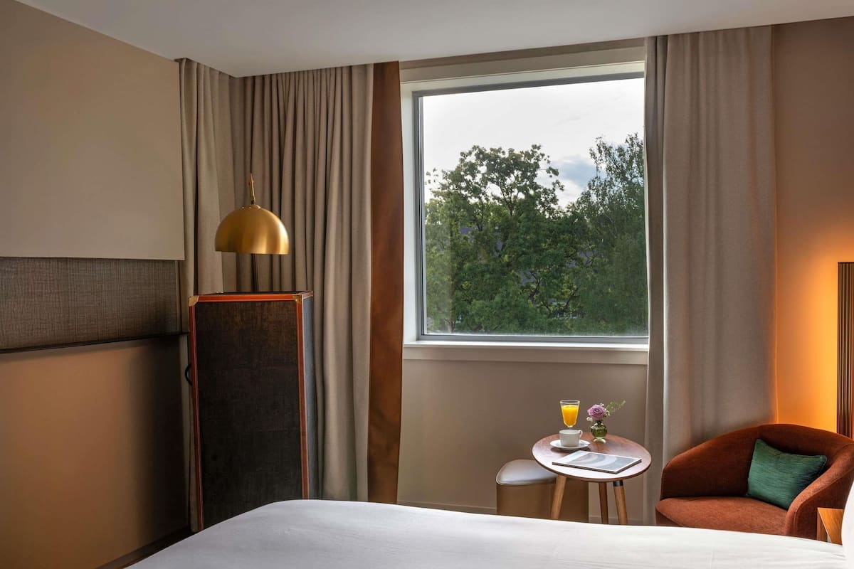 Ein Hotelzimmer mit einem Bett, einem kleinen Tisch mit Getränk und Blume, einem Sessel und einem Fenster mit Blick auf Bäume.