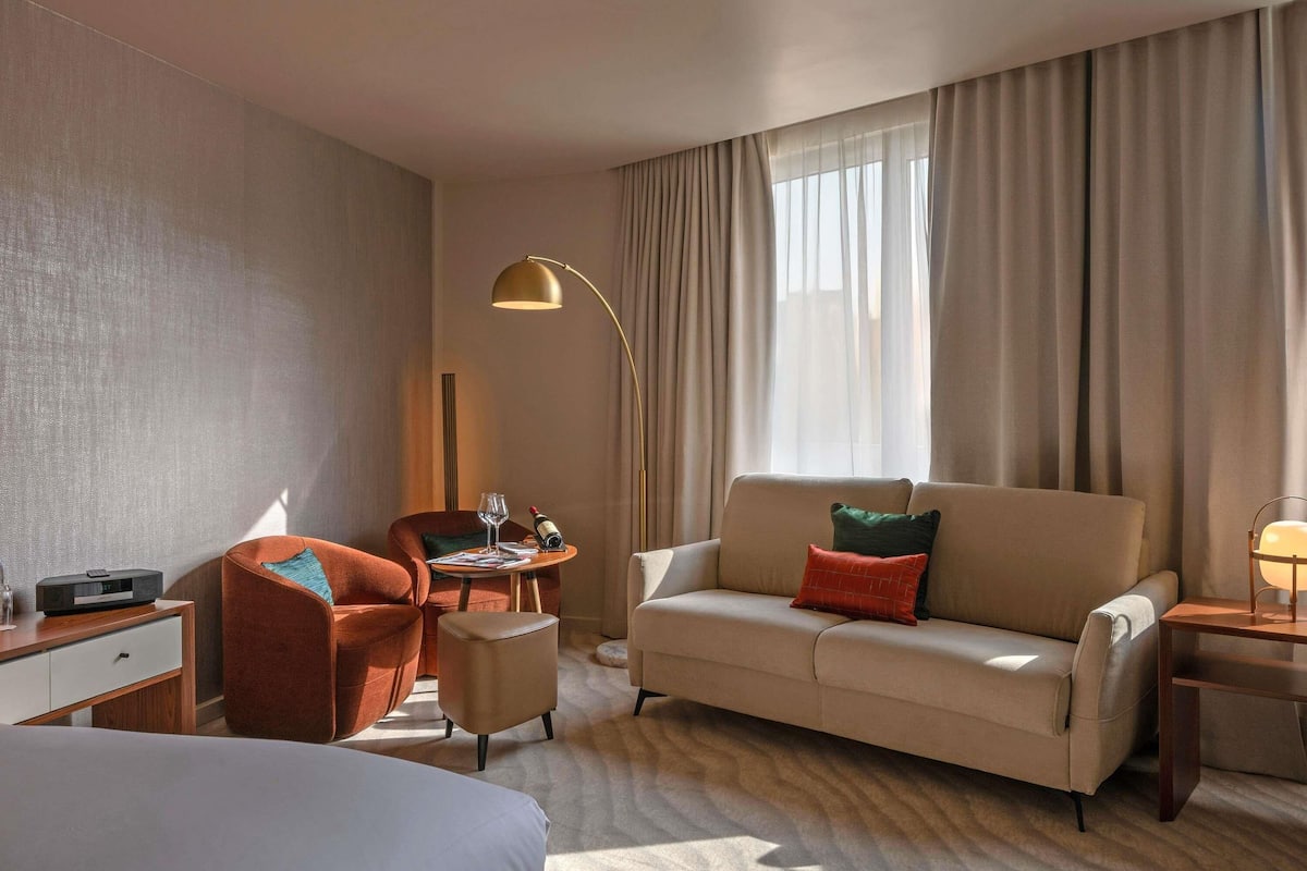 Una habitación de hotel con cama, sofá, escritorio con lámpara y ventana con cortinas.