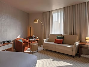 Une chambre d’hôtel avec un lit, un canapé, un bureau avec une lampe, une chaise et une fenêtre avec des rideaux.