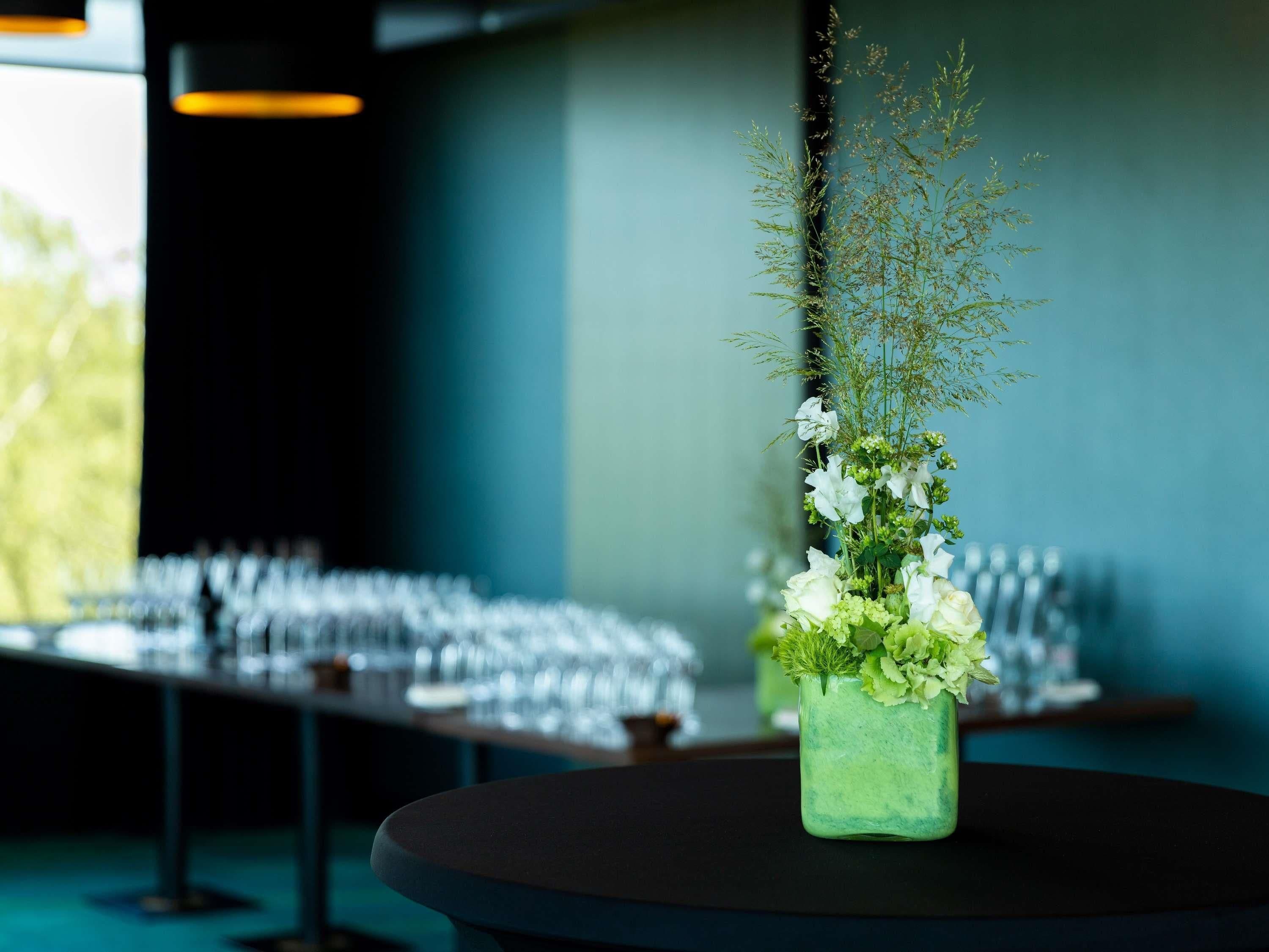 A table with a floral arrangement in a green vase, a row of chairs, and a window in the background.