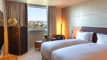 A hotel room with two beds, a desk, a chair, a lamp, a small table with a plant, and a view of the city through the window.
