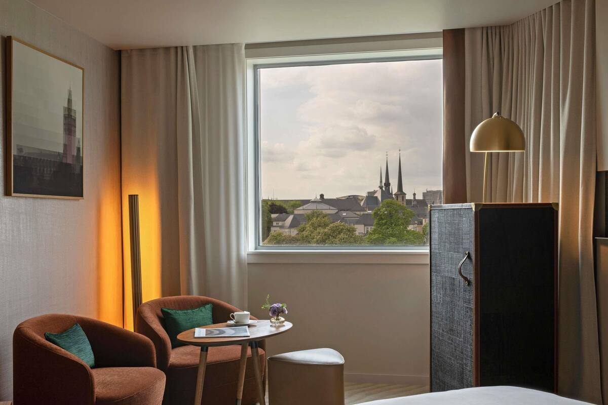Ein Hotelzimmer mit Bett, einem Sessel, einem kleinen Tisch mit Tasse und Blumenvase, einer Lampe und einem Fenster mit Blick auf die Stadt.