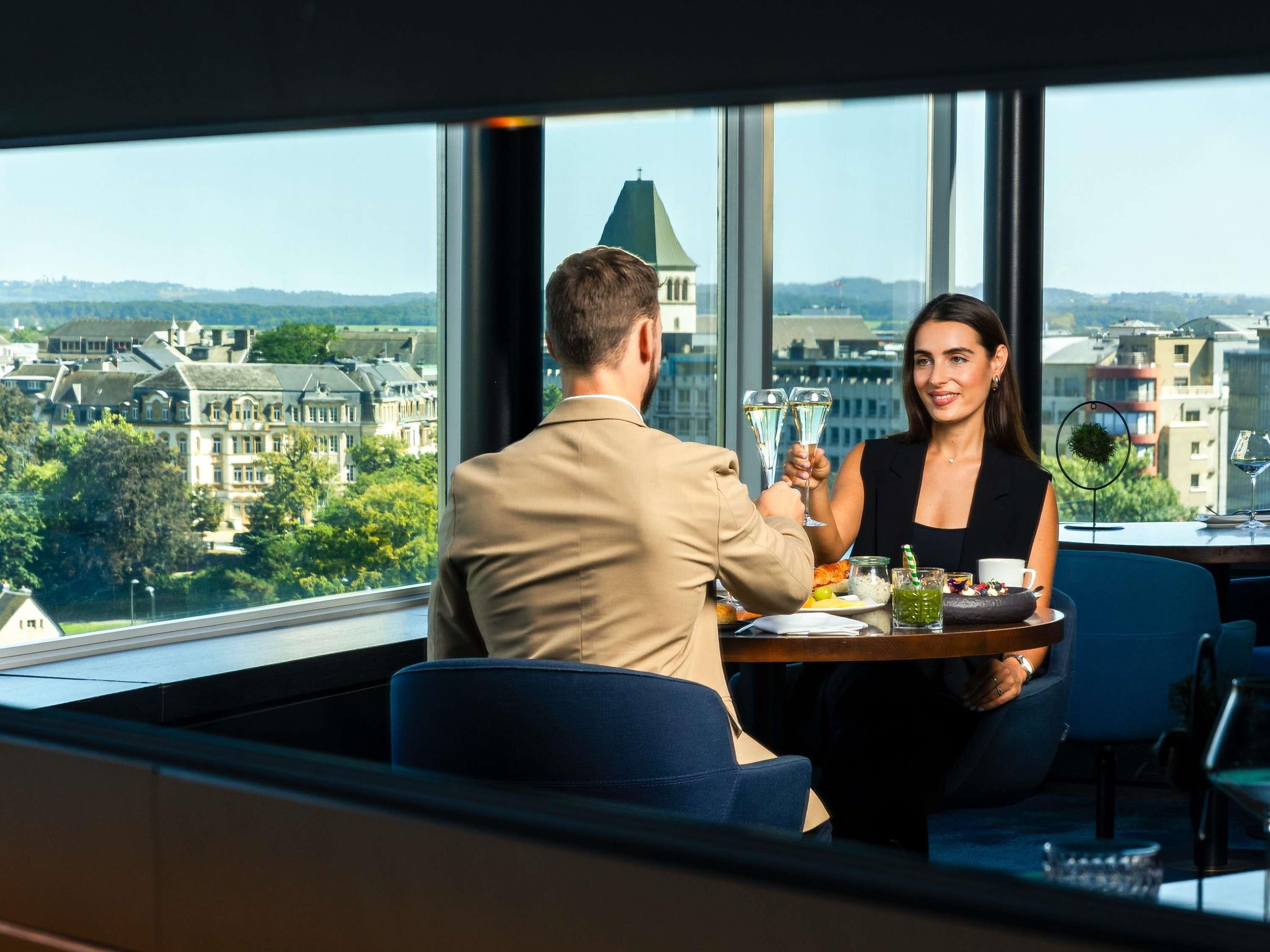 Ein Mann und eine Frau sitzen an einem Tisch mit Blick auf die Stadt, ein Tablett mit Speisen und Getränken, und ein Fenster mit Blick auf Gebäude und Bäume.