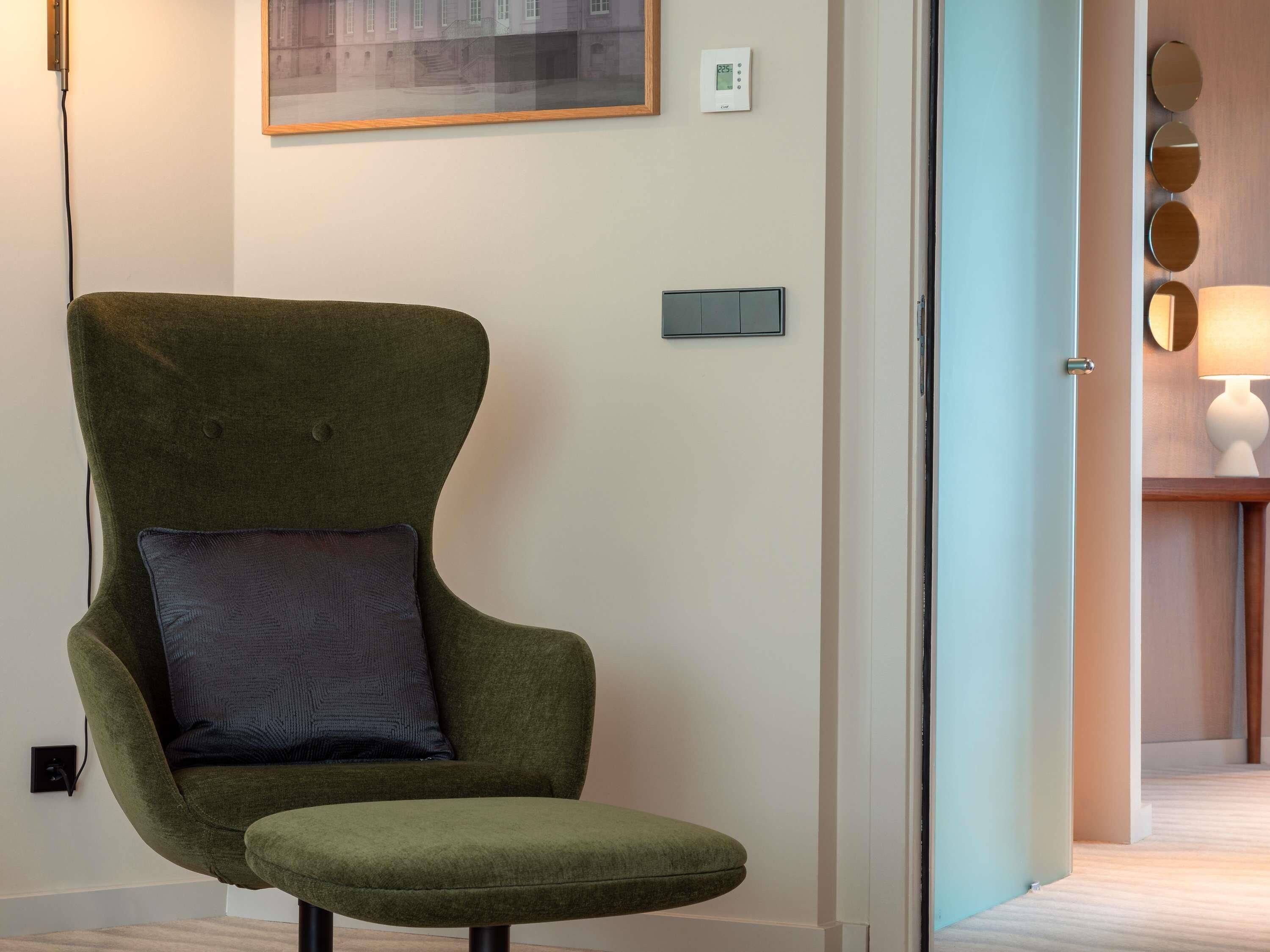 Ein grüner Sessel mit Kissen, ein gerahmtes Bild an der Wand, ein Thermostat und ein Spiegel mit runden Rahmen.