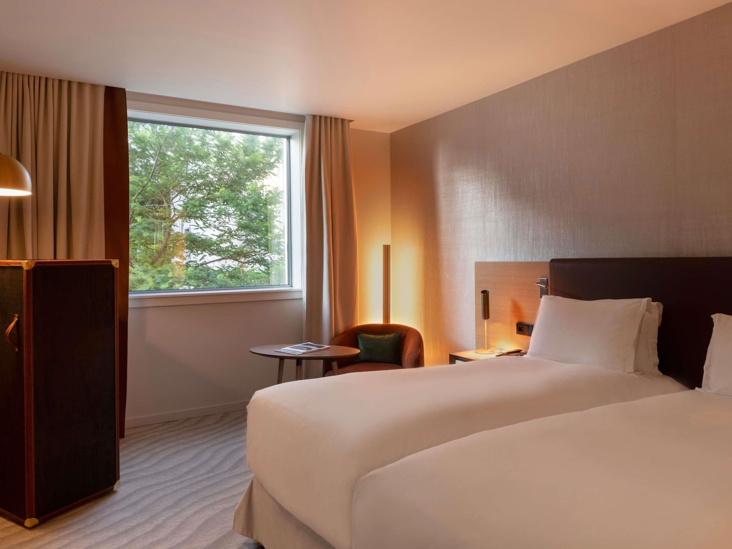 Habitación de hotel con una cama grande, un escritorio, una silla, una lámpara y una ventana con cortinas.