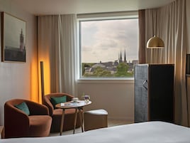 A hotel room with a bed, a chair, a small table with a cup and a vase, a lamp, and a window with a view of buildings and a church.